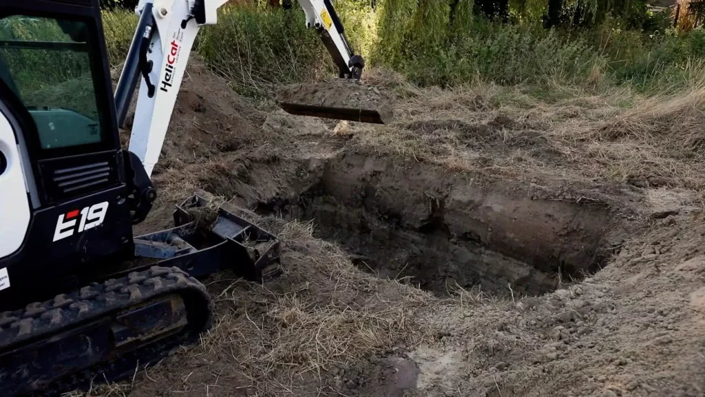 Mini-pelleteuse creusant une fosse rectangulaire dans un terrain herbeux