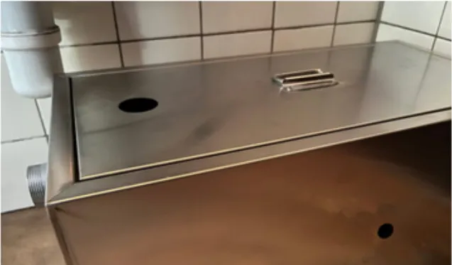 Dégraisseur en inox posé contre un mur carrelé blanc, près d’un tuyau d’évacuation
