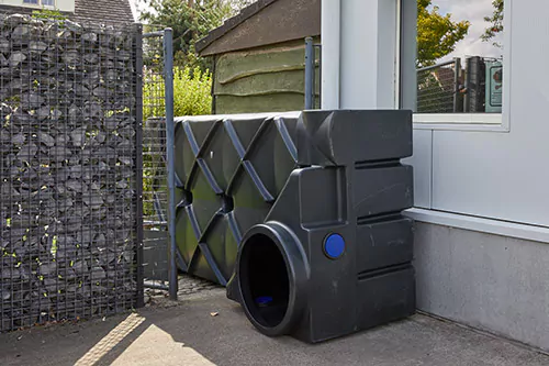 Citerne plate noire posée sur la tranche dans un passage étroit, entre une clôture en gabions et un bâtiment