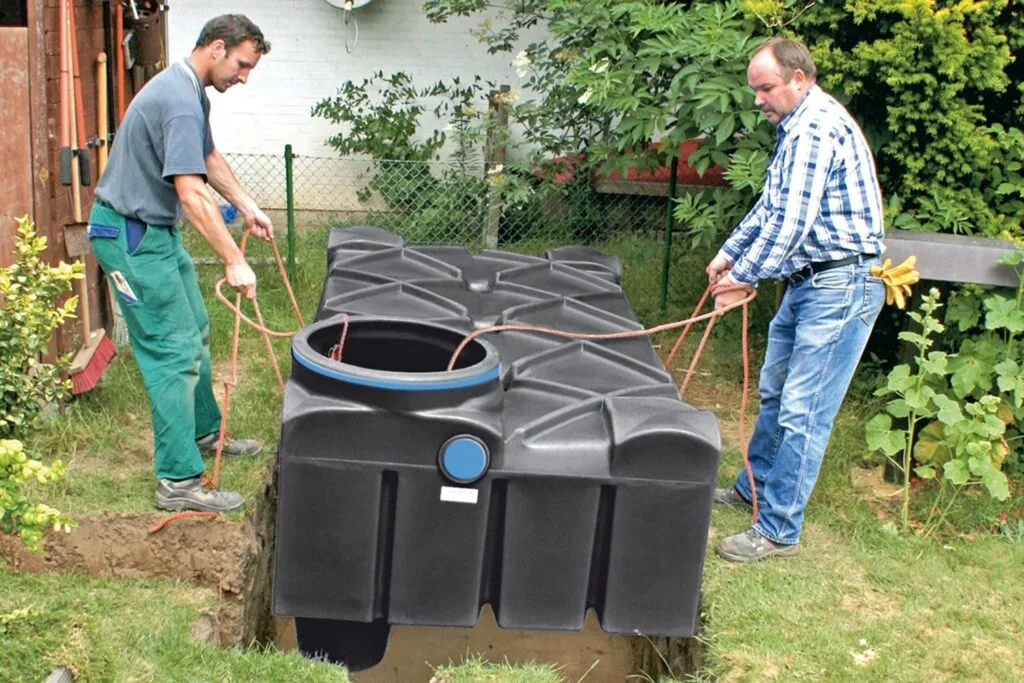 Deux hommes descendent à l’aide de cordes une fosse septique plate noire dans un trou creusé dans un jardin